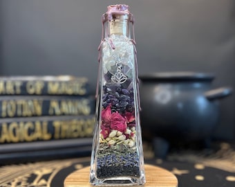 May include: A clear glass bottle filled with various crystals, dried flowers, and herbs. The bottle has a cork stopper and a silver charm hanging from it. The bottle is sitting on a wooden coaster.