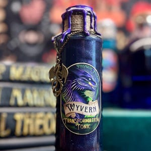 May include: A dark blue potion bottle with a purple wax seal and a bronze dragon charm. The label reads "Wyvern Transformation Tonic" with a dragon illustration. The bottle has a glittery, shimmering effect, suggesting a magical theme.