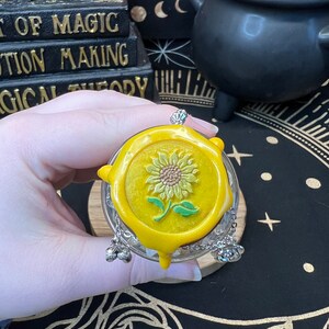 May include: A yellow glass bottle with a yellow wax seal featuring a sunflower design. The bottle has a silver chain with three silver charms attached to the top.