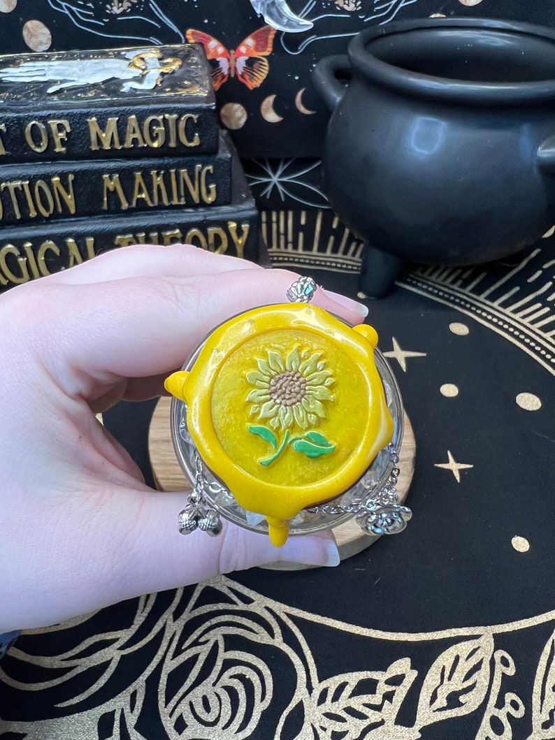 May include: A yellow glass bottle with a yellow wax seal featuring a sunflower design. The bottle has a silver chain with three silver charms attached to the top.