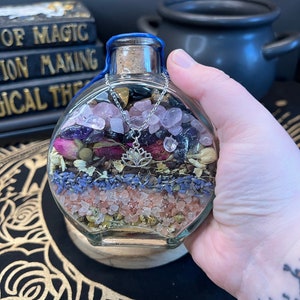 May include: A clear glass bottle filled with various crystals, dried flowers, and herbs. The bottle has a blue lid and a silver charm hanging from the top. The bottle is sitting on a black and gold patterned surface.