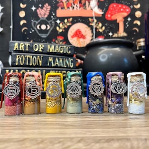 May include: Seven small glass bottles with cork stoppers and silver charms. Each bottle is filled with different colored herbs and crystals. The bottles are arranged in a row on a wooden surface. The bottles are sealed with wax in different colors, including red, orange, yellow, green, blue, purple, and white.