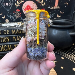 May include: A small glass bottle filled with dried flowers and herbs, sealed with a yellow wax. The bottle is held in a hand.