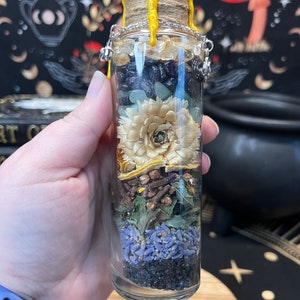 May include: A glass bottle filled with various dried herbs, flowers, and crystals. The bottle is sealed with a cork and has a yellow wax seal. The bottle is hanging from a silver chain.