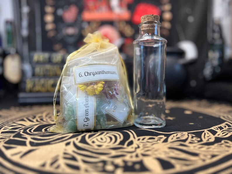 May include: A clear glass bottle with a cork stopper, next to a yellow organza bag filled with dried flowers and green crystals. The bag has labels that read "6. Chrysanthemum" and "7. Green Aventurine".
