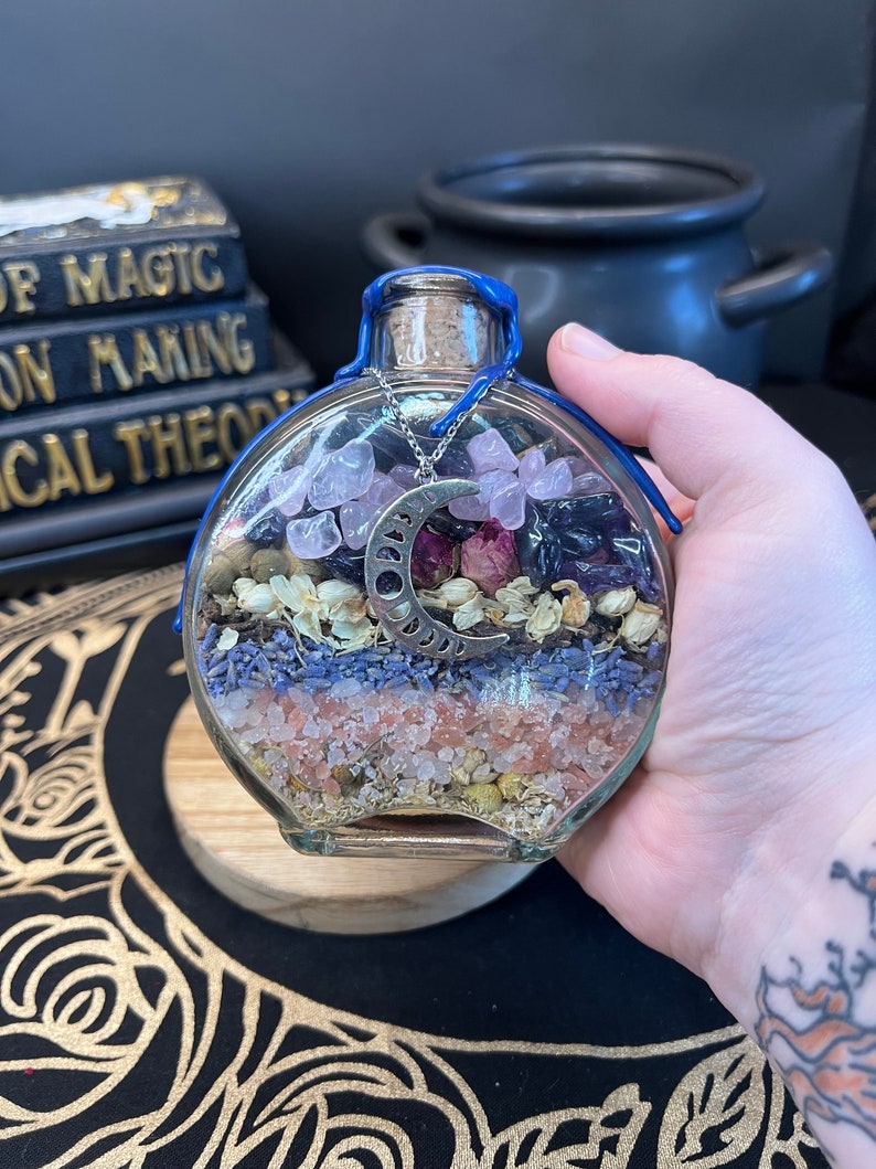 May include: A glass bottle filled with layers of pink Himalayan salt, dried lavender, chamomile, and rose petals. The bottle is topped with a silver crescent moon charm and a blue ribbon.
