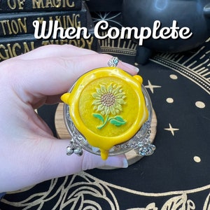 May include: A yellow glass jar with a yellow wax seal featuring a green sunflower design. The jar has a silver chain with three silver bells attached.
