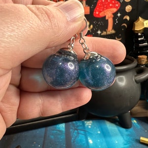 May include: A pair of silver hoop earrings with blue glitter resin balls. The balls have a dark blue base with silver glitter throughout.