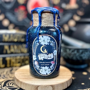 May include: A dark blue glass bottle filled with glitter, labeled "Fairy Dust." The bottle is sealed with twine and blue wax. The label features a crescent moon, fairy, and floral design. The bottle sits on a small wooden stand.