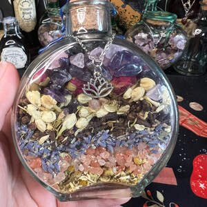 May include: A clear glass bottle filled with dried herbs, flowers, and crystals. The bottle has a cork stopper and a silver chain with a lotus charm hanging from it. The bottle is sitting on a black and red patterned surface.