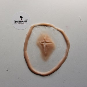 Puede incluir: Primer plano de un molde de silicona de color carne con un diseño de cicatriz en forma de cruz. El molde está sobre una superficie blanca.