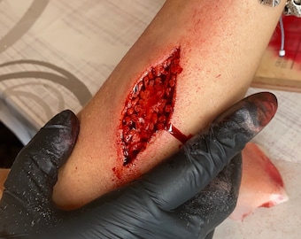 May include: A close-up of a fake wound on a person's arm. The wound is a deep gash with red and white makeup to simulate blood and flesh.