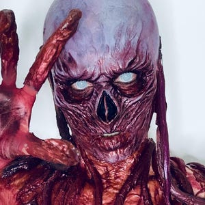 Peut inclure: Un masque grotesque et détaillé avec une tête violet pâle et des traits du visage charnus exposés. Le masque a des yeux blancs brillants, un nez sombre et une expression menaçante. Des vrilles rouge foncé, semblables à des veines, s'étendent du visage.