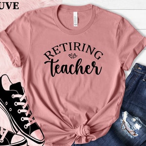 以下が含まれることがあります： 「Retiring Teacher」の黒い文字がプリントされた、モーヴ色のTシャツ。青いジーンズと白と黒のスニーカーが写っています。Tシャツはウエストで結ばれています。