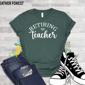 以下が含まれることがあります： 「Retiring Teacher」の白文字がプリントされた濃い緑色のTシャツ。青いジーンズと白と黒のスニーカーと組み合わせています。「Heather Forest」の文字が上にあります。