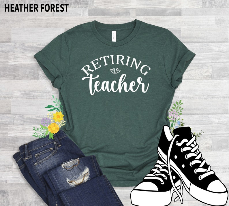 以下が含まれることがあります： 「Retiring Teacher」の白文字がプリントされた濃い緑色のTシャツ。青いジーンズと白と黒のスニーカーと組み合わせています。「Heather Forest」の文字が上にあります。