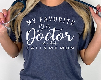 Könnte beinhalten: Ein kurzärmeliges T-Shirt in Heather Navy mit weißem Text, der MY FAVORITE Doctor CALLS ME MOM lautet, mit einem kleinen Stethoskop-Symbol über dem Wort Doctor und einer Herzschlaglinie, die darunter zentriert dargestellt ist.