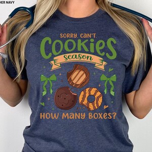 May include: Heather navy t-shirt with the text "Sorry, Can't. Cookies Season. How Many Boxes?" in green and orange. The design includes illustrations of Girl Scout cookies.