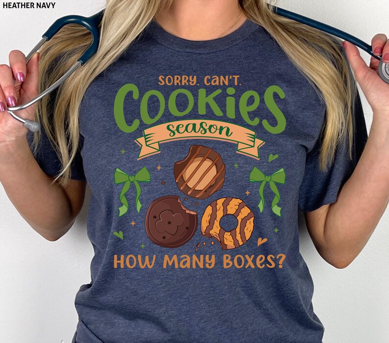 May include: Heather navy t-shirt with the text "Sorry, Can't. Cookies Season. How Many Boxes?" in green and orange. The design includes illustrations of Girl Scout cookies.