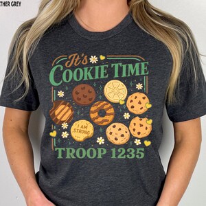 Puede incluir: Camiseta gris oscuro con el texto "It's Cookie Time" e ilustraciones de galletas. La camiseta tambi&eacute;n presenta el texto "I AM STRONG" y "TROOP 1235". El dise&ntilde;o incluye margaritas y corazones.