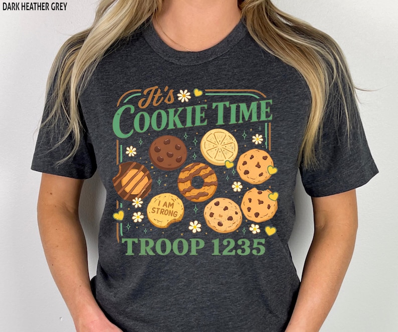 Puede incluir: Camiseta gris oscuro con el texto "It's Cookie Time" e ilustraciones de galletas. La camiseta tambi&eacute;n presenta el texto "I AM STRONG" y "TROOP 1235". El dise&ntilde;o incluye margaritas y corazones.