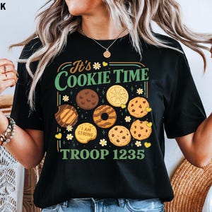 Puede incluir: Camiseta negra con un gr&aacute;fico colorido que presenta varias galletas y el texto "It's Cookie Time" y "Troop 1235". El dise&ntilde;o incluye ilustraciones de chispas de chocolate, lim&oacute;n y otras galletas.