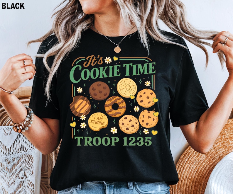 Puede incluir: Camiseta negra con un gr&aacute;fico colorido que presenta varias galletas y el texto "It's Cookie Time" y "Troop 1235". El dise&ntilde;o incluye ilustraciones de chispas de chocolate, lim&oacute;n y otras galletas.