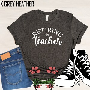 以下が含まれることがあります： 「Retiring Teacher」の文字が白で書かれたダークグレーヘザーTシャツ。画像には、青いジーンズと白と黒のスニーカーも含まれています。