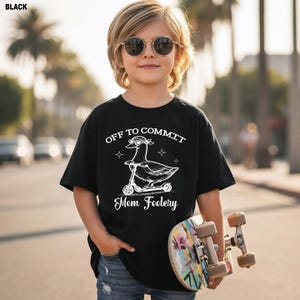 May include: A black t-shirt featuring a white graphic of a duck wearing heart-shaped sunglasses while riding a scooter. The text above the duck reads OFF TO COMMIT and the text below the duck reads Mom Foolery. The shirt has short sleeves.