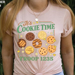 Puede incluir: Camiseta melocot&oacute;n claro con el texto "It's Cookie Time" en verde y naranja. La camiseta presenta ilustraciones de varias galletas y el texto "TROOP 1235".