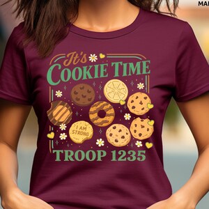 Puede incluir: Camiseta granate con el texto "It's Cookie Time" en verde y amarillo, con ilustraciones de varias galletas. La camiseta tambi&eacute;n muestra "I AM STRONG" y "TROOP 1235".