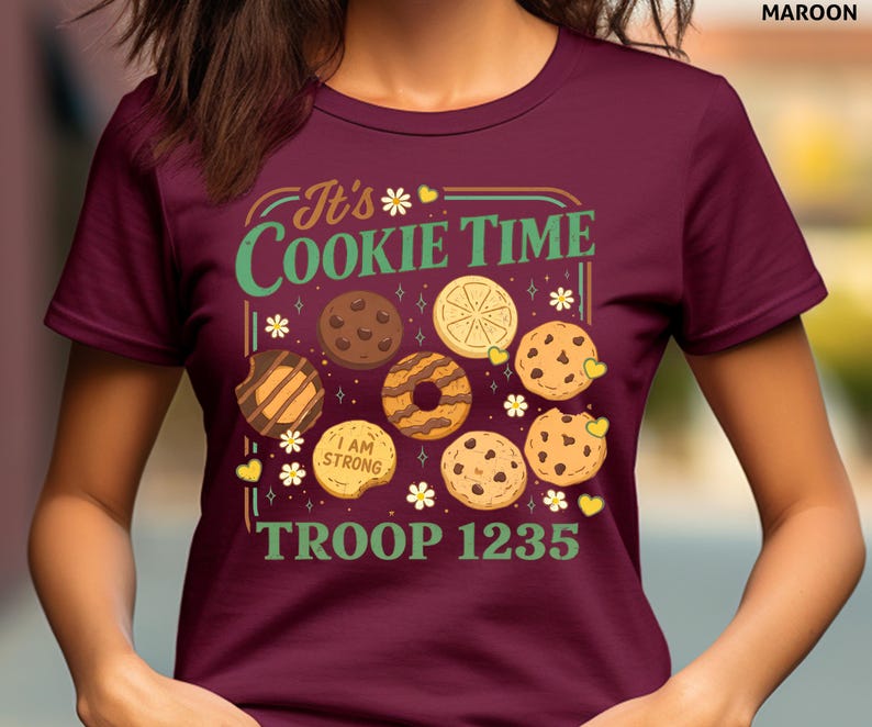 Puede incluir: Camiseta granate con el texto "It's Cookie Time" en verde y amarillo, con ilustraciones de varias galletas. La camiseta tambi&eacute;n muestra "I AM STRONG" y "TROOP 1235".