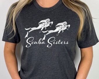 May include: A dark heather grey short-sleeve t-shirt featuring a white graphic of two scuba divers swimming. Below the divers, the text Scuba Sisters is printed in a white cursive script font. The shirt has a crew neckline and a standard fit.