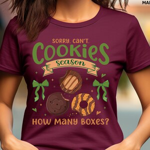 May include: Maroon t-shirt with the text "Sorry, Can't. Cookies Season" in green and tan. The shirt features illustrations of cookies and green bows. The phrase "How Many Boxes?" is printed at the bottom.