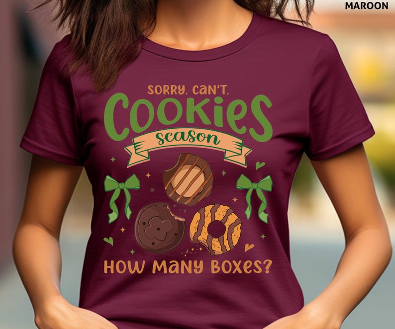 May include: Maroon t-shirt with the text "Sorry, Can't. Cookies Season" in green and tan. The shirt features illustrations of cookies and green bows. The phrase "How Many Boxes?" is printed at the bottom.