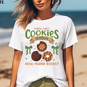 May include: White t-shirt with the text "SORRY, CAN'T. Cookies season. HOW MANY BOXES?" in green and brown. The shirt features illustrations of cookies and green bows.