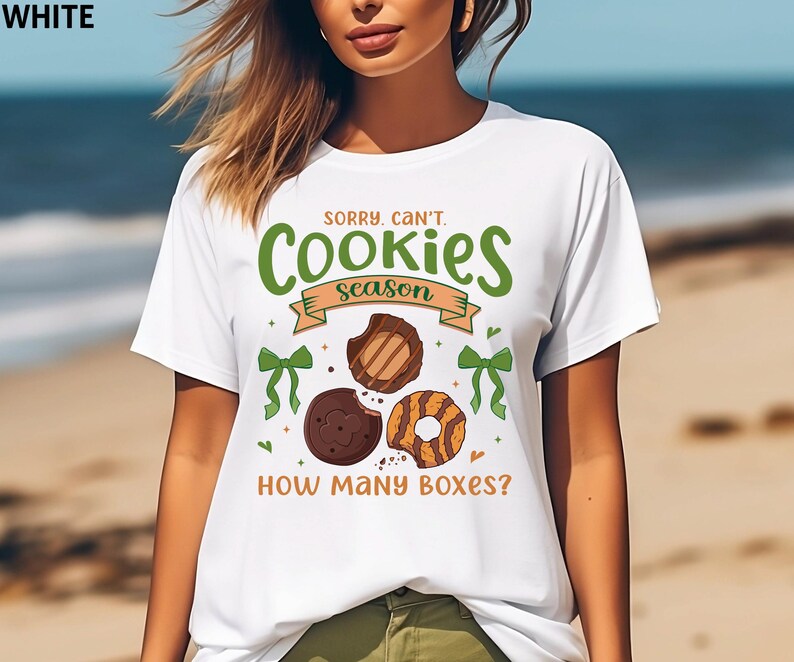 May include: White t-shirt with the text "SORRY, CAN'T. Cookies season. HOW MANY BOXES?" in green and brown. The shirt features illustrations of cookies and green bows.