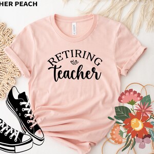 以下が含まれることがあります： 「Retiring Teacher」の黒い文字が書かれた、ヘザーピーチ色のTシャツ。 黒と白のスニーカーと花の配置でスタイリングされています。 画像の左上隅には「HEATHER PEACH」という文字があります。