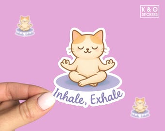 May include: A sticker featuring a cartoon cat in a yoga pose, with closed eyes and a serene expression. The cat is light brown with white markings, sitting on a blue cloud with the words "Inhale, Exhale". Additional smaller stickers are in the background.