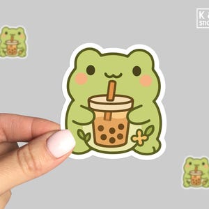 May include: A green frog sticker holding a boba tea. The frog has rosy cheeks and a white outline. Two smaller frog stickers are also visible. The sticker has the text "K & O STICKERS".