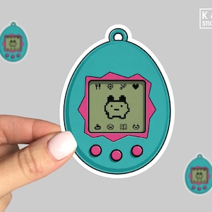 May include: A teal egg-shaped digital pet sticker with a pink jagged border around the screen. The screen displays a pixelated creature and various icons. Three pink circular buttons are positioned below the screen, and a small loop is at the top.