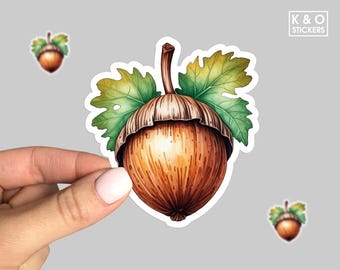 Puede incluir: Una ilustración de acuarela de una bellota con una tapa marrón y una nuez marrón claro. La bellota está rodeada de hojas verdes.