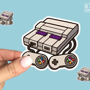 May include: A sticker featuring a cartoon illustration of a Super Nintendo Entertainment System (SNES) console with two controllers. The console is gray and white with purple accents. The controllers are gray with colorful buttons.