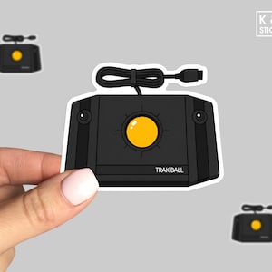 May include: Black Trak-Ball sticker with a yellow trackball and a black cord. The sticker has the text "TRAK-BALL" on the front. The sticker is held by a hand. There are two smaller stickers of the same design in the background.