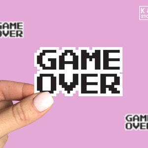 May include: A white sticker with black pixelated text that reads "GAME OVER". The sticker is being held by a hand against a pink background.