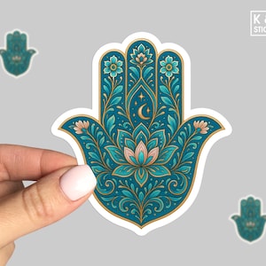 May include: A teal and gold Hamsa hand sticker with floral and celestial details. The sticker features a lotus flower, crescent moon, and stars. The hand is held by a person. The sticker is from K & O Stickers.