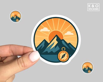 May include: A round sticker featuring a mountain range, a rising sun, and a compass. The design uses shades of orange, yellow, and teal. The sticker is held by a hand. The text "K & O STICKERS" is in the upper right corner.