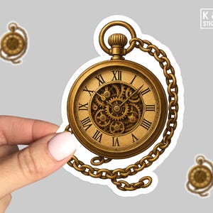 May include: A gold-colored pocket watch sticker with Roman numerals and visible gears. The watch is attached to a chain. Two smaller pocket watch stickers are also visible. The sticker has a white border and is held by a hand.