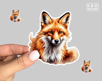 May include: A sticker featuring a detailed watercolor illustration of a fox with orange and white fur. The fox is the main subject, with two smaller fox stickers in the background. The sticker has a white border and the text "K & O STICKERS".