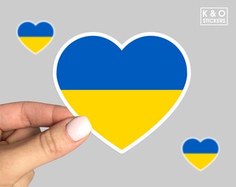 May include: A heart-shaped sticker featuring the blue and yellow horizontal stripes of the Ukrainian flag. The design includes a white border. In the top right corner, a small white square contains the text K & O STICKERS.